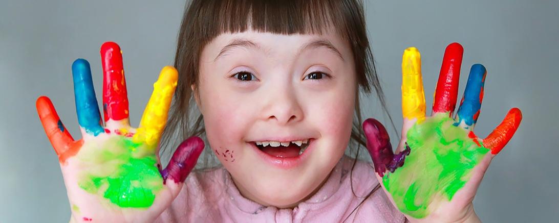 Das Bild zeigt ein Kind mit Trisomie 21, welches die Hände voller Farbe hat. 