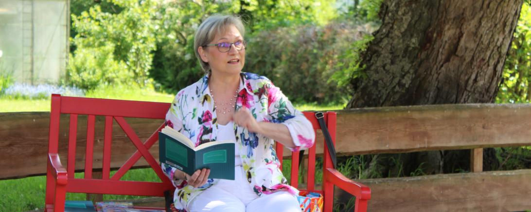 Eine Autorin sitzt auf einer roten Bank und liest vor. 
