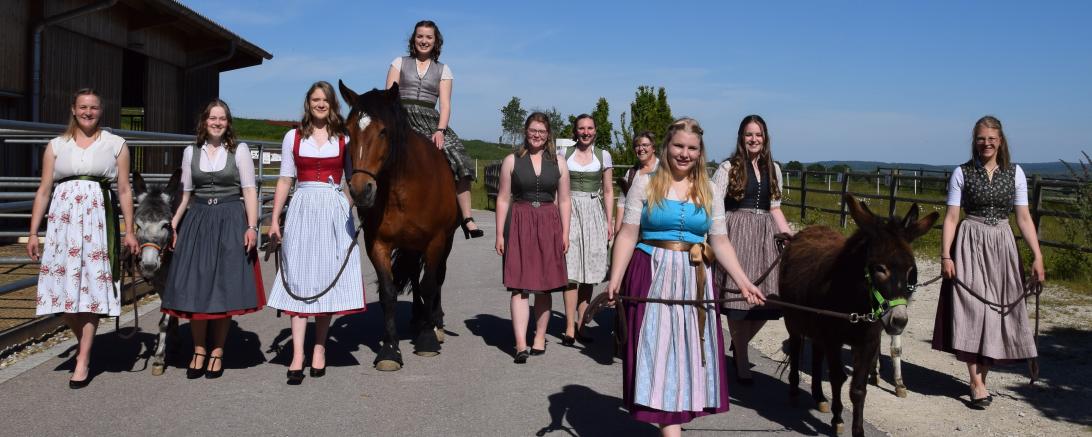 Zehn Frauen im Dirndl laufen auf Straße, eine reitet auf einem Pferd, dabei zwei Esel