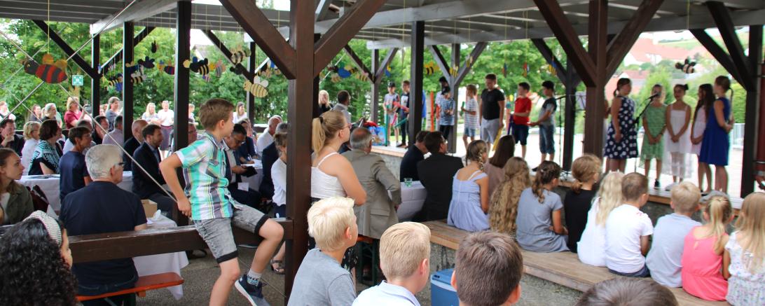 Kinder singen ein Lied vor Publikum unter einer hölzernen Überdachung