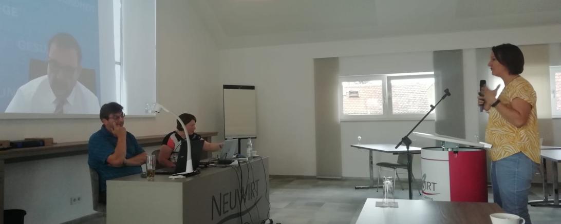 Leinwand mit Klaus Holetschek, davor Mann und Frau an einem Podium, rechts Frau an Mikrofonständer