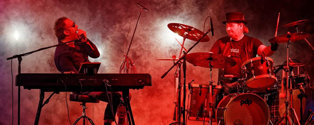 Keyboarder und Schlagzeuger auf Bühne in rot-lila Licht und Nebel