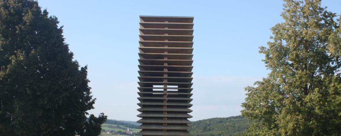 Turm aus Holzplatten zwischen Bäumen, im Hintergrund Häuser