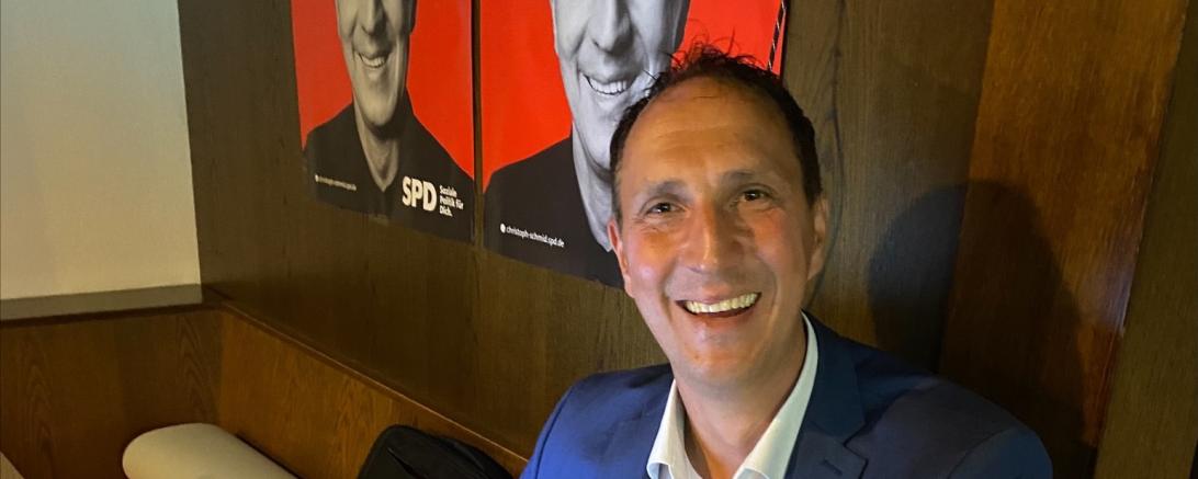 Christoph Schmid sitzt lächelnd an Laptop, im Hintergrund zwei seiner Wahlplakate