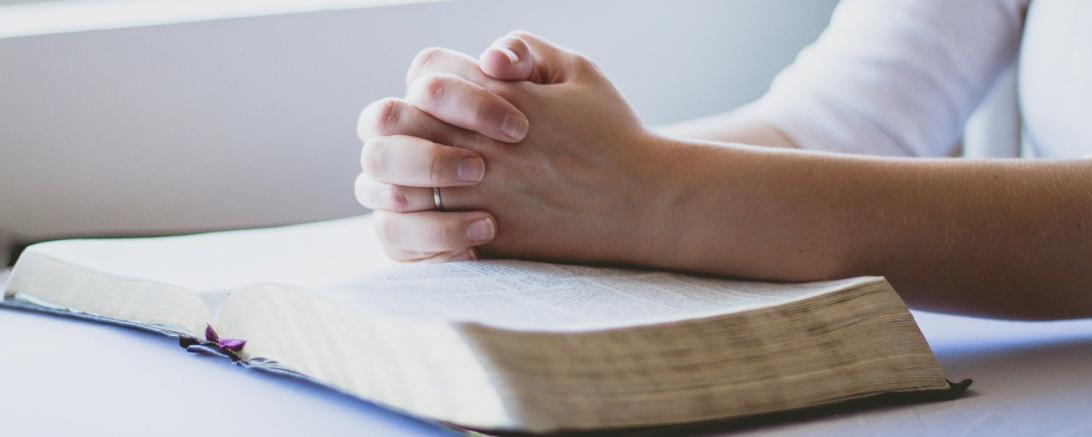 Auf dem Bild sieht man Hände, zum Gebet gefaltet, auf einer offenen Bibel ruhend