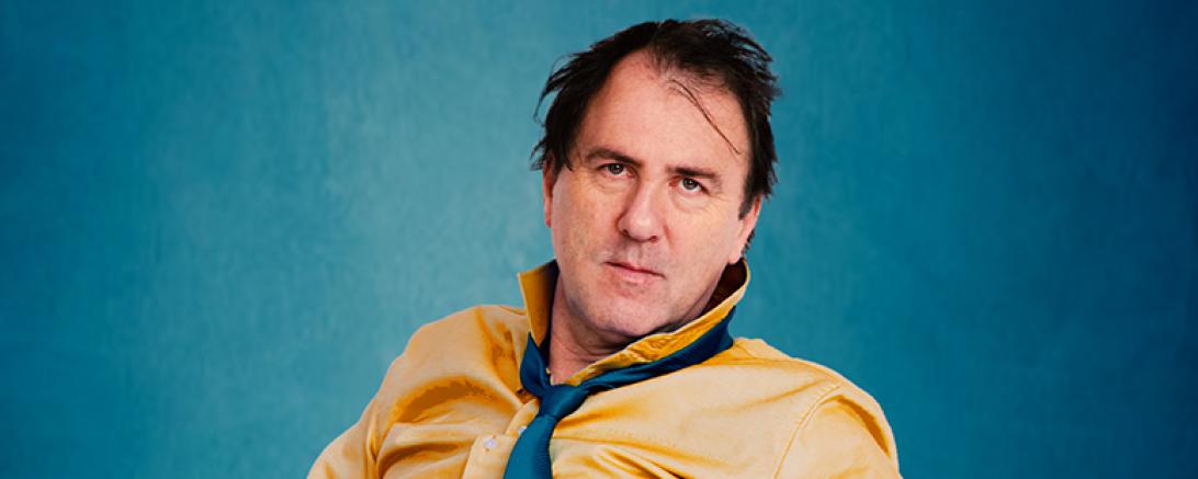 Mann im gelben Hemd mit blauer Krawatte sitzt vor blauem Hintergrund