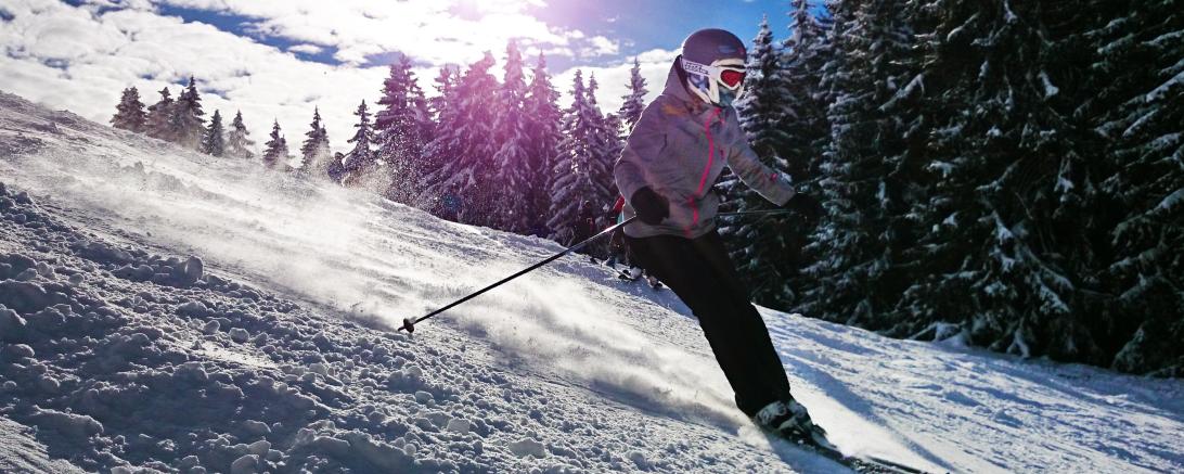 Skifahrerin fährt Berg hinunter, dahinter Bäume und tiefstehende Sonne