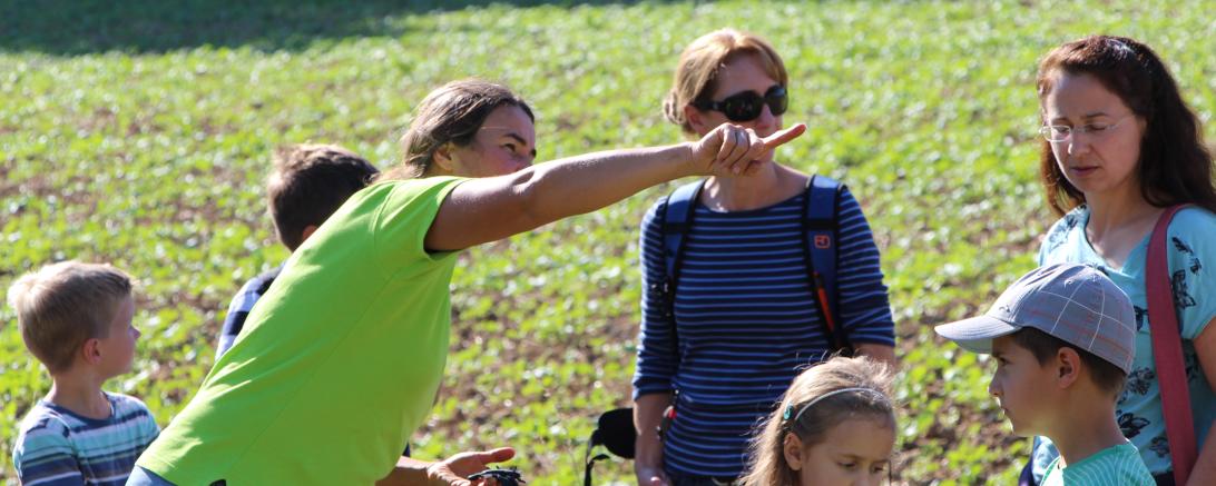 Zu sehen ist eine Frau, welche vor einer Gruppe von Menschen mit dem Finger auf etwas in der Natur zeigt.