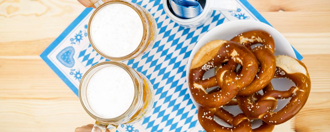 Zu sehen sind zwei Bierkrüge, welche von zwei Händen zum Anstoßen gehalten werden, daneben steht ein Teller mit Brezeln. 