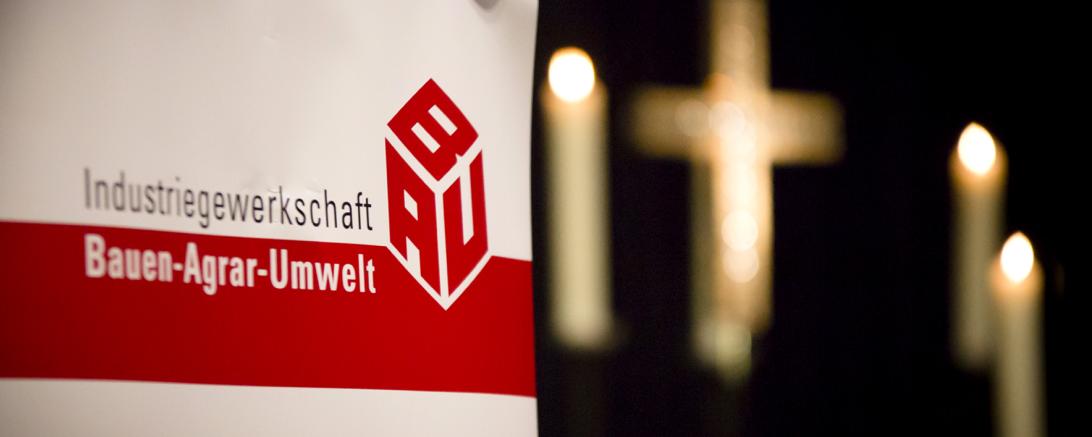 Zu sehen ist ein Plakat der IG BAU vor einem Kreuz und drei Kerzen. 