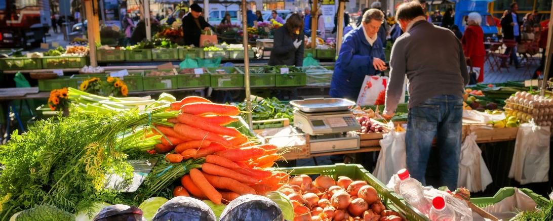 Zu sehen ist ein Marktstand mit Gemüse, bei dem gerade jemand einkauft. 