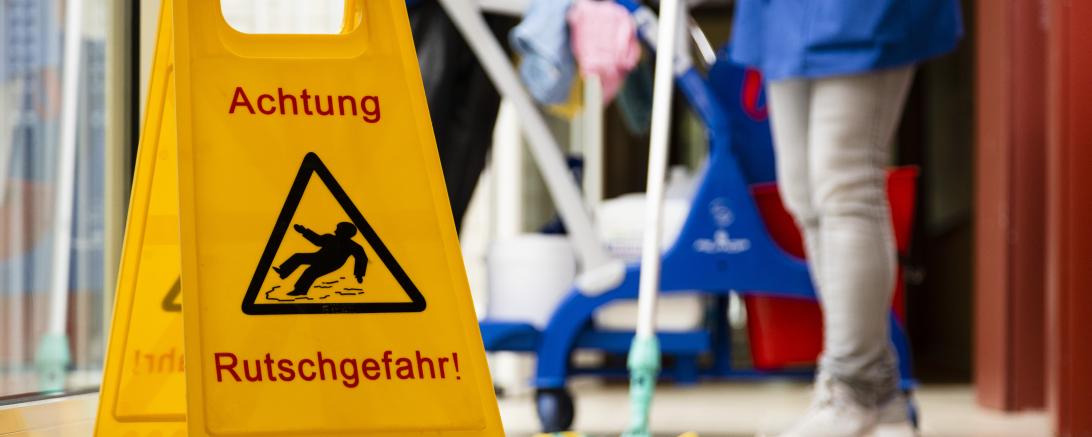 Zu sehen ist ein Schild mit der Aufschrift "Achtung Rutschgefahr" und eine Reinigungskraft im Hintergrund. 