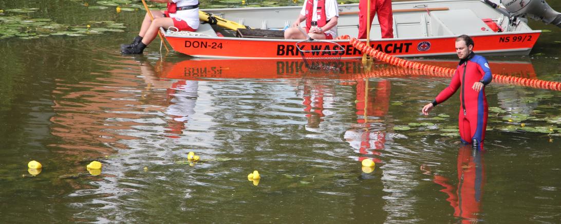 Zu sehen sind mehrere Menschen in einem Boot und Plastikenten im Wasser. 