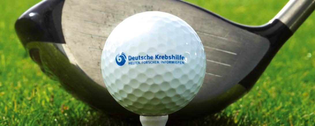 Zu sehen ist ein Golfball mit der Aufgschrift "Deutsche Krebshilfe" vor einem Golfschläger. 