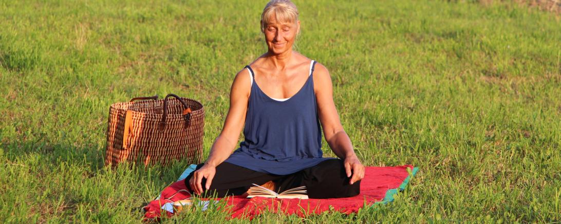 Zu sehen ist eine Frau auf einer Wiese im Schneidersitz. 