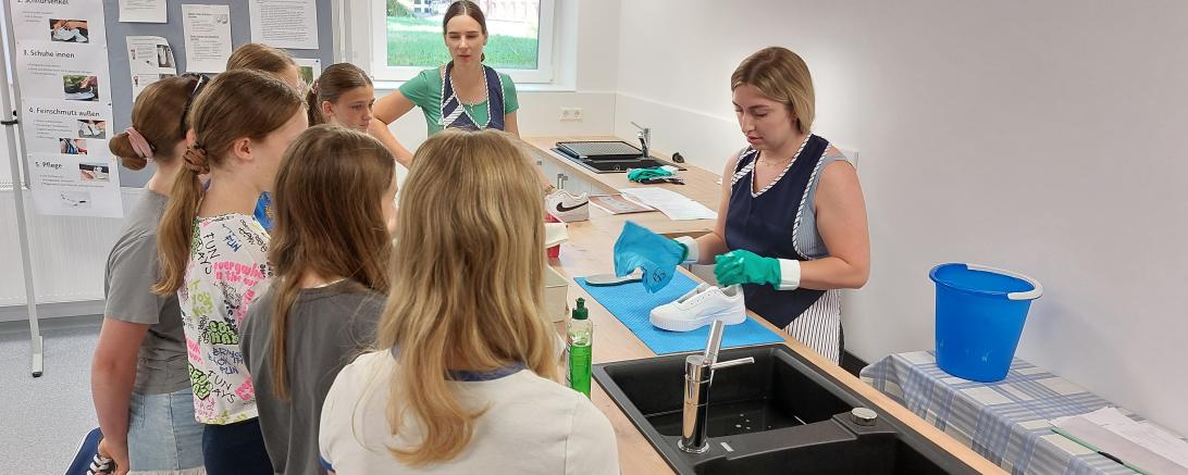 Das Foto zeigt die Fachschülerinnen Maria Sinning (li.) und Anna-Maria Wiedemann (re.) mit einer Gruppe Schülerinnen. Anna-Maria Wiedemann führt vor, welche Schritte bei der Schuhreinigung wichtig sind.