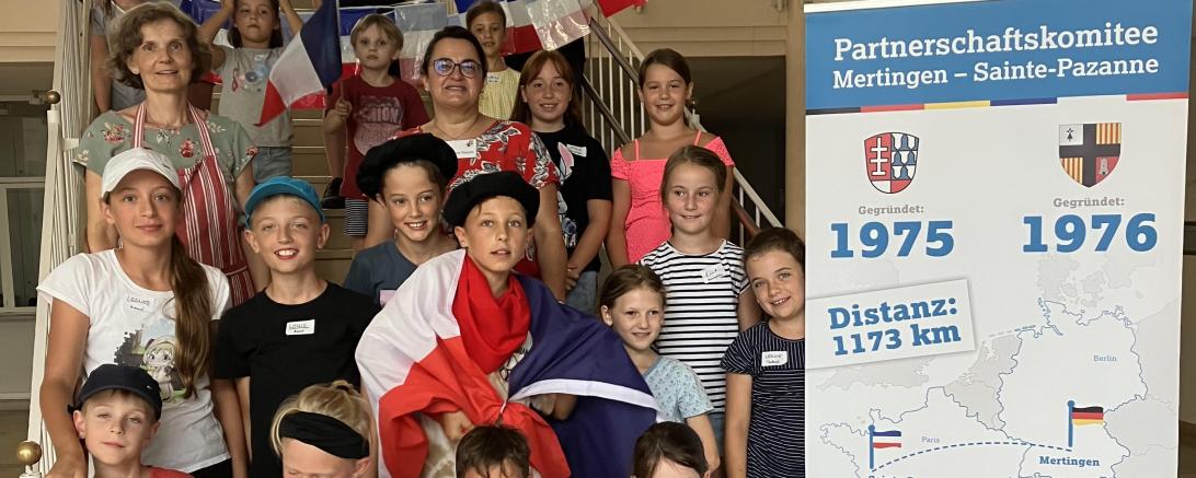 Marita Braun und Christine Riepold, Vorsitzende des Partnerschaftskomitees, mit den Kindern des Ferienprogramms in der Aula der Grundschule Mertingen.