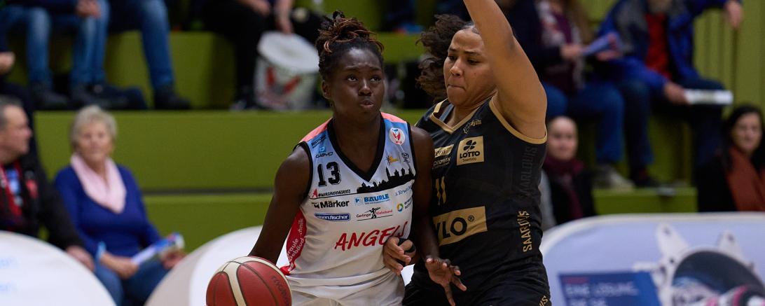 Zwei Basketballspielerinnen, die linke Person hält den Ball unten auf der linken Seite mit einer Hand und die rechte Person hält die rechte Hand nach oben und versucht den Ball abzuwehren.