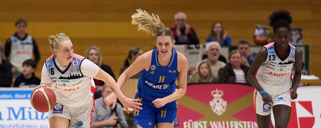 Drei Basketballerinnen, zwei kämpfen um einen Ball, eine hat ein Blaues Trikot an, die andere hat ein Weißes Trikot an