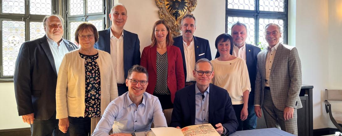 Michael Müller Trägt sich ins Goldene Buch der Stadt Nördlingen ein, Er sitzt an einem Tisch der eine Blaue Tischdecke drüber hat. Hinter ihn sind Personen.