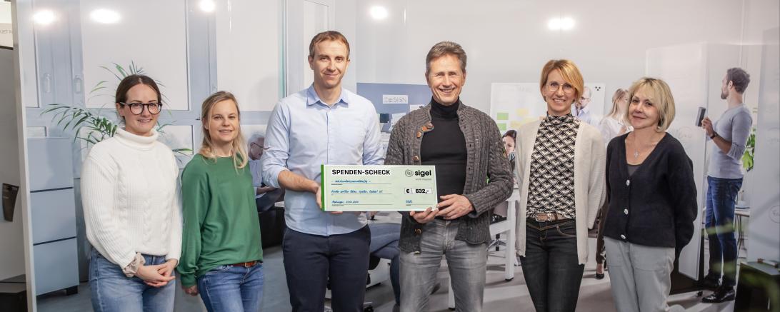 Zwei Menschen halten einen Check und neben den beiden Stehen links und rechts auf jeder Seite zwei weitere Menschen.