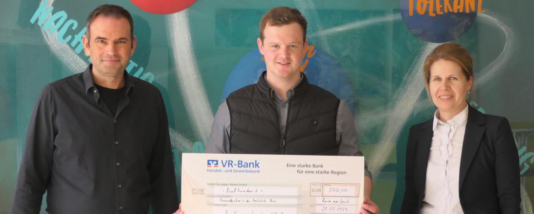 Ein Mann, der einen großen Check der hält und in der Mitte steht, neben ihm steht links und rechts eine Person