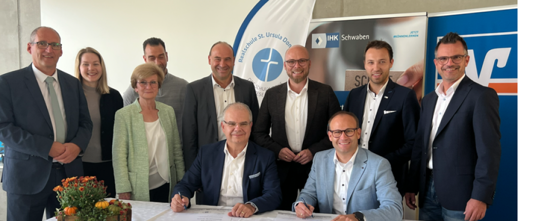 Kooperation zwischen der Raiffeisen-Volksbank Donauwörth eG und der Realschule St. Ursula.  Vorstandssprecher Michael Kruck (sitzend, links) und Schulleiter Tobias Freißler (sitzend, rechts)  unterzeichnen die Verträge im Beisein von Vertretern aus Politik und Wirtschaft.  
