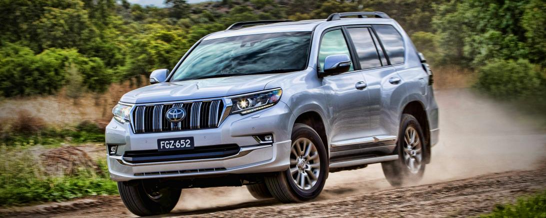 Silberner Toyota Land Cruiser Prado auf einem erdigen und staubigen Feldweg zwischen Bäumen.
