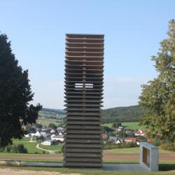 Turm aus Holzplatten zwischen Bäumen, im Hintergrund Häuser